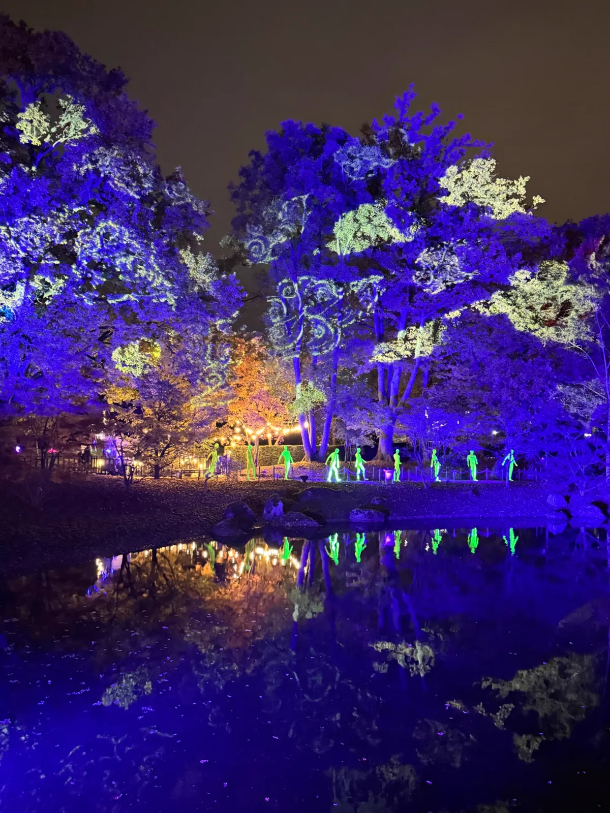 Blauw verlichte bomen met geprojecteerde kumo-wolkenmotieven en lichtgevende figuren aan de vijver